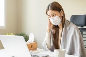 Sick asian young woman wearing mask wrapped blanket have headache have fever, flu and using laptop computer consulting with doctor online, telehealth at home. Health care people on virus seasonal. photo