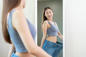 Shape slender, thin waist, attractive slim asian young woman, beautiful girl hand show shape her weight loss, wearing in big, large or over size jeans and looking into mirror. People body fit healthy. photo