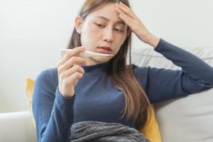 enfermo, influenza asiático joven mujer, niña mano en cheque dolor de cabeza, tener un fiebre, gripe y cheque termómetro medida cuerpo temperatura, sensación enfermedad sentado en sofá cama a hogar. salud cuidado persona en virus. foto