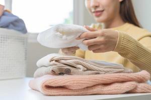Housewife, asian young woman hand in many folding freshly shirts, sweaters or dress on desk, table after washing clean clothes and drying, making household working in room at home. Laundry and maid. photo