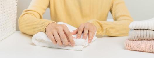 Housewife, asian young woman hand in many folding freshly shirts, sweaters or dress on desk, table after washing clean clothes and drying, making household working in room at home. Laundry and maid. photo