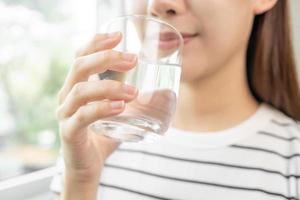 Happy beautiful, asian young woman, girl drinking, sip fresh glass of water for hydration of body, holding transparent glass in her hand, thirsty at home. Health care, healthy lifestyle concept.