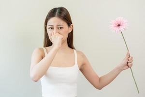 Allergic rhinitis symptom of odor pollen flowering, portrait asian young woman hand in sneeze, blowing runny nose after smell, smelly from bloom flower, holding away pink flora on white background. photo