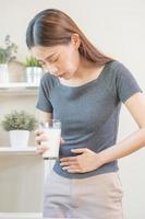 dolor, alergia asiático joven mujer, niña mano en barriga sufrir después bebida vaso de leche, infeliz y malo estómago dolor. lactosa intolerancia y lechería comida , salud problema concepto. foto