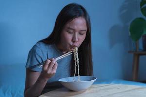 Diet, templtation. Alone asian young student woman, girl using chopsticks eating instant ramen, noodles in bowl on table in bedroom in overtime night, late time at home. photo