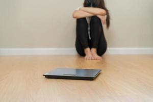 Deprimido y estrés, triste asiático joven mujer, niña cubrir cara con mano, sentado en piso, peso en escala en frente de su. ganancia perder peso y exceso de peso gordo. personas dieta, insalubre, infeliz concepto foto