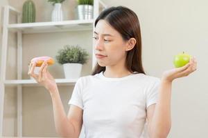 dieta, dieta asiático joven mujer o niña sonrisa, confuso elegir, elección verde manzana o rosado dulce rosquilla, a hogar, comer comida para bueno saludable, salud cuando hambriento. cerca arriba hembra peso pérdida persona. foto