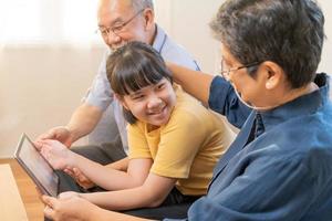 Happy family asian aged grandfather, grandmother have fun, smiling parent, girl using touchpad watching funny video call, play games and take photo selfie enjoy weekend together in living room at home