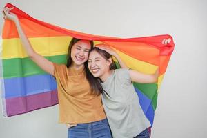 Happy lesbian, beautiful asian young two women, girl gay, couple love moment spending good time together, holding or waving lgbt rainbow, pride flag on isolated white wall background together at home. photo