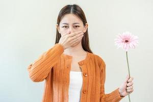 Allergic rhinitis symptom of odor pollen flowering, portrait asian young woman hand in sneeze, blowing runny nose after smell, smelly from bloom flower, holding away pink flora on white background. photo