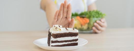 dieta, dieta asiático joven mujer utilizando mano empujar afuera, negar chocolate pastel o dulce gusto, postre alimento, escoger comer verde ensalada vegetales cuando hambriento. nutricionista de saludable, nutrición de peso pérdida. foto