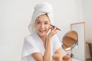 contento belleza blogger concepto, hermosa asiático joven mujer, niña en bata de baño maquillaje cara por aplicando negro delineador de ojos, mano en poniendo en ojo mirando a el espejo. personas Mira con natural Moda estilo foto