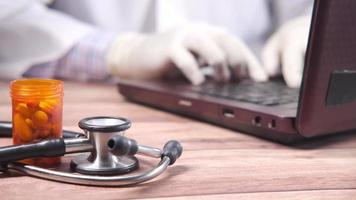 Doctor working on laptop with pills container and stethoscope on table video