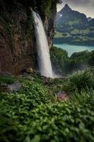 A mighty waterfall flowing out of a mountain surrounded by green grass, a person in a yellow raincoat standing in front of it photo