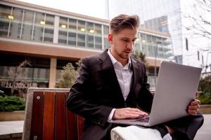 un joven chico en un traje trabajos en un computadora en el parque, mira dentro un computadora foto