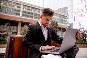 un joven chico en un traje trabajos en un computadora en el parque, mira dentro un computadora foto