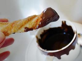 A hand holding a churros stick dipped with hot thick chocolate, ready to eat, Spanish coffee shop, blurred white cup background photo