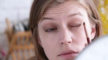 Daily Makeup. Natural beauty. Young woman without makeup looking at the mirror brushing her eyebrows with eyebrow pencil, yellow mimosa flowers an the background video