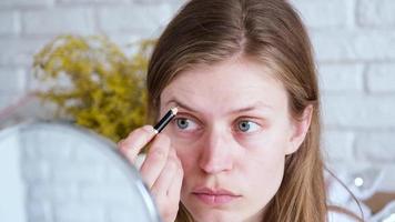 Daily Makeup. Natural beauty. Young woman without makeup looking at the mirror brushing her eyebrows with eyebrow pencil, yellow mimosa flowers an the background video