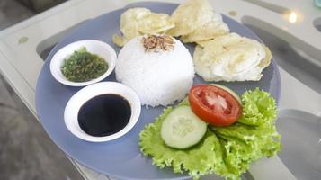 A plate containing of a white rice, lettuce, a slice of tomato and cucumber, crackers, soy sauce and crushed green chilies photo