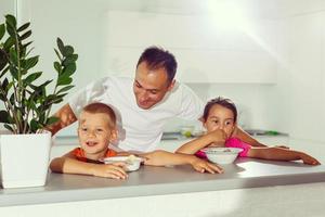 Happy young family, father with two children, adorable toddler girl and funny messy boy having healthy breakfast eating fruit and dairy, sitting in a white sunny kitchen with window photo