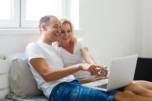 Young couple relaxing in bed. Happy affectionate young couple relaxing in bed enjoying themselves laughing in amusement with their laptop computer. Interracial couple, Asian woman, Caucasian man.. photo