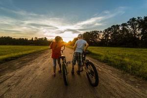 niños montando uno bicicleta juntos en soleado verano noche. sentado en bicicleta estante. familia de dos personas disfrutando de viaje en escénico campo terminado puesta de sol cielo antecedentes. foto