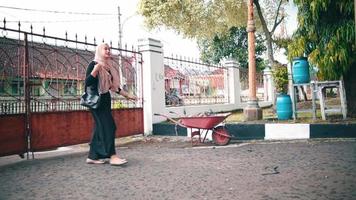 a Muslim woman with a pink headscarf and a black shirt walks in front of her house video