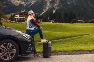 Young woman sitting in the car trunk with suitcases, mountains in the background photo