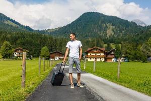 Traveler man walking with suitcase in the street in the mountains. Travel concept. photo