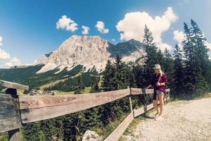 Rear view of a female hiker walking on a trail in the Alps near Ehrwald, Tyrol, Austria. Landscape with grass, trees, rocky mountains and blue sky. photo