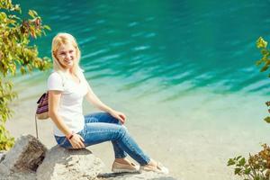 back view of blonde girl in hat with backpack standing near lake with sunlight photo