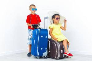 Children travelling - boy holding a camera, girl in sunglasses and fashionable hat sit on a suicase. Isolated, white background photo