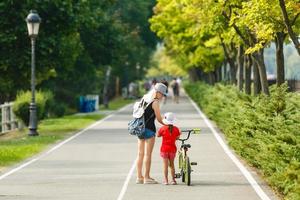 Beautiful and happy young mother teaching her cute daughter to ride a bicycle. Both smiling and looking at each other. Summer park in background photo