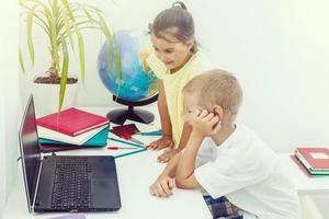 Education at home concept - Cute little Indian Asian kids studying on study table with pile of books, educational globe, laptop computer, coffee mug etc photo