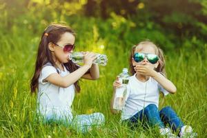 dos pequeño muchachas son Bebiendo un botella de agua en el parque. concepto de pureza, ecológico y biológico producto, amor para naturaleza foto