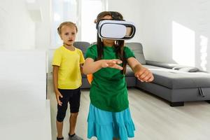 Lifestyle shot of an amazed two little kids using a virtual reality goggles with mouth open shocked seated in the living room at home. photo