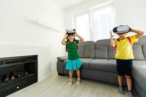 Lifestyle shot of an amazed two little kids using a virtual reality goggles with mouth open shocked seated in the living room at home. photo