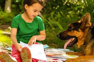 Little girl with his dog sitting on green grass and read e-book. photo
