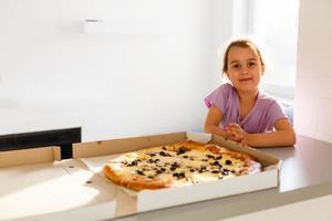 Charming happy young girl laugh and biting off big slice of fresh made pizza. She sit at white chair in Provence style interior, smile and enjoy sunny day and yummy meal. She has long blonde hair. photo