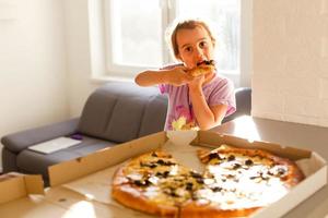 Portrait of cute hungry happy smiling little girl eating tasty pizza sitting by dinner table photo