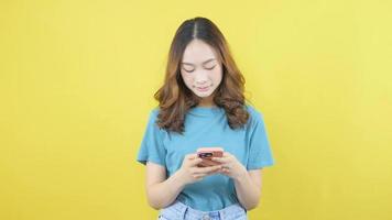 souriant asiatique femme en utilisant mobile téléphone, texte discuter, permanent dans T-shirt plus de Jaune Contexte copie espace. video