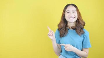 vídeo de asiático mujer sonriente mientras señalando izquierda y pulgares arriba presentando producto en pie en amarillo antecedentes. video