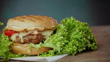 Close up of Big Burger lies on craft white paper against wooden table. A Juicy green Salad leaf and a red Tomato lie near the Burger video