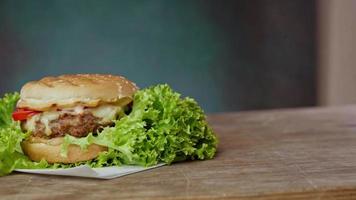 dichtbij omhoog van groot hamburger leugens Aan ambacht wit papier tegen houten tafel. een sappig groen salade blad en een rood tomaat liggen in de buurt de hamburger. 4k resolutie video