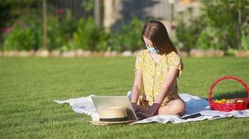 Young girl wearing face mask on a blanket in the grass with laptop and basket of fruit video