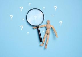 A wooden toy puppet holds a magnifying glass and question marks on a blue background, concept of answering questions and gaining knowledge photo