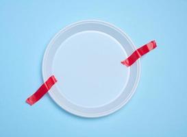 Empty white plastic plate attached with red adhesive tape on a blue background, a concept of refusing plastic tableware and recycling materials, zero waste photo