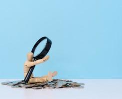A wooden mannequin is holding a stack of coins against the background of a magnifying glass. The concept of income growth photo