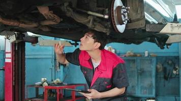 Professional young Asian male motor mechanic inspects undercarriage of electric car lifted by forklift jack for repair at garage, automotive maintenance service works industry occupation business. video
